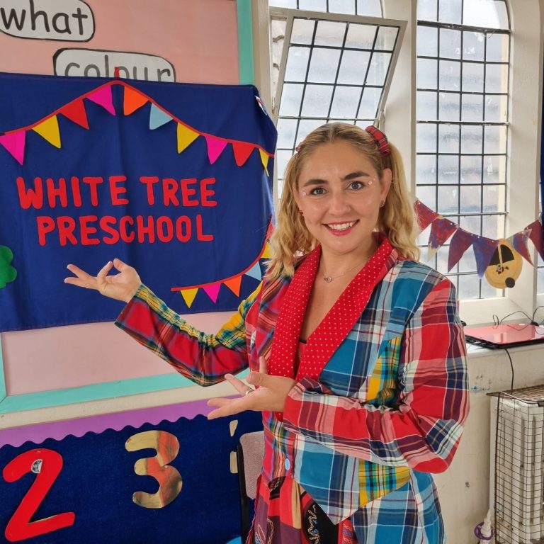 Children laughing during a fun magic and games show Woman in a colourful outfit points to a sign reading "White Tree Preschool".