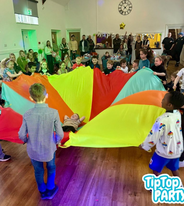 Birthday party fun with dancing, games and laughter. Children and adults playing with a large, brightly coloured parachute in a lively room.
