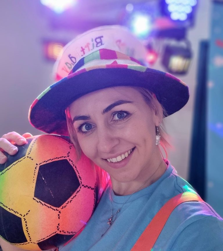 Colourful kids’ disco party with music and dancing entertainment. Smiling woman holding a colourful soccer ball, wearing a festive hat and bright clothing.